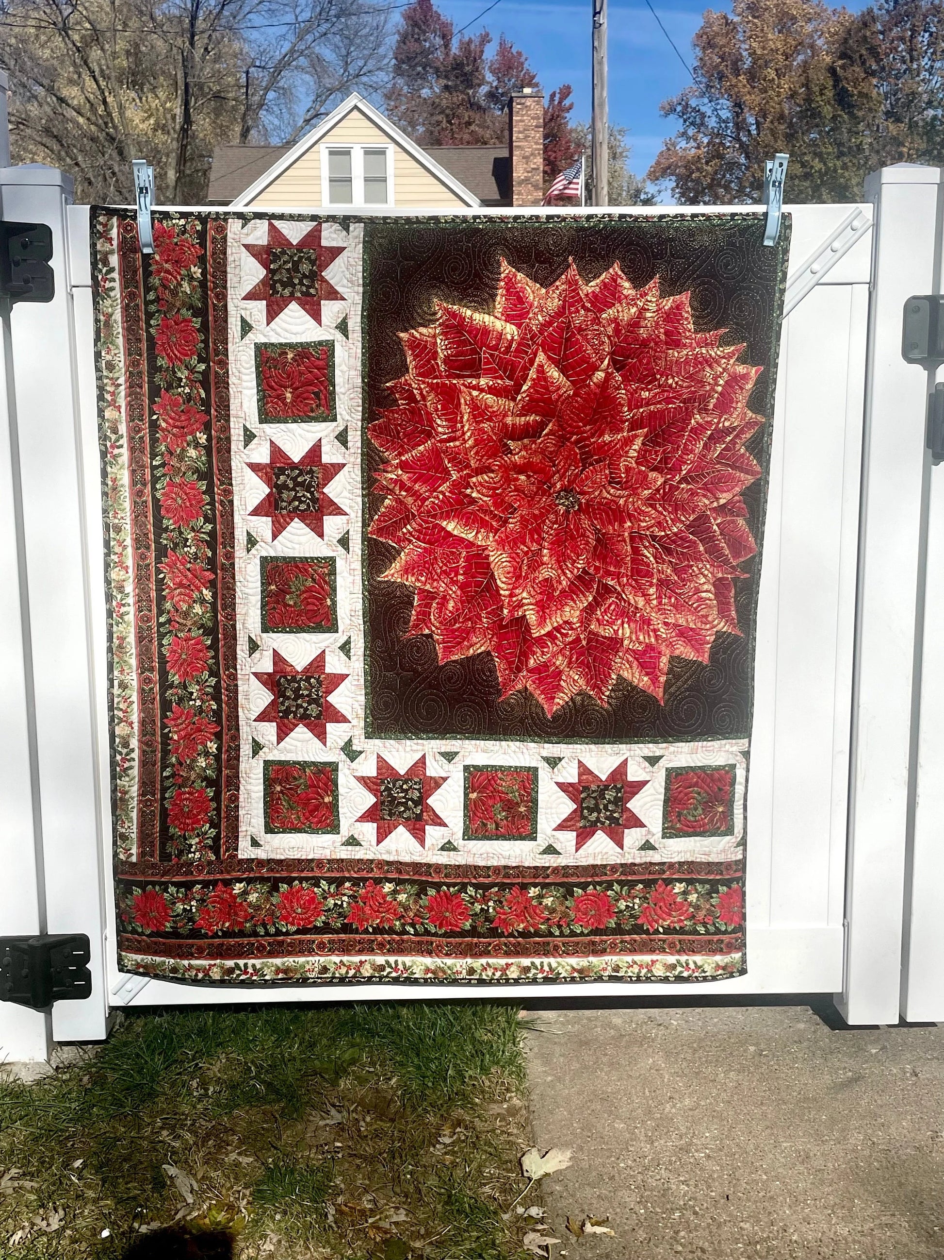 Poinsettia wall hanging quilt. There is a large red and gold poinsettia flower in the top right corner on a black background. There is a border on the left and bottom with red, black, and white quilted stars. There is an additional border on the left and bottom with a row of red poinsettia flowers and greenery. It is an appropriate size for a quilted wall hanging or a lap blanket quilt.